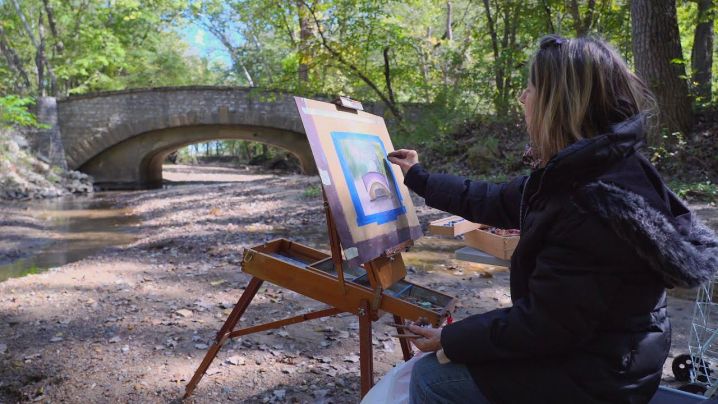 A female artist draws in pastels near a stone arch bridge and stream.