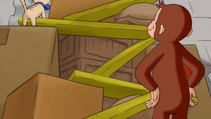 Curious George observes a cat climbing up wood planks connecting levels of stacked crates.