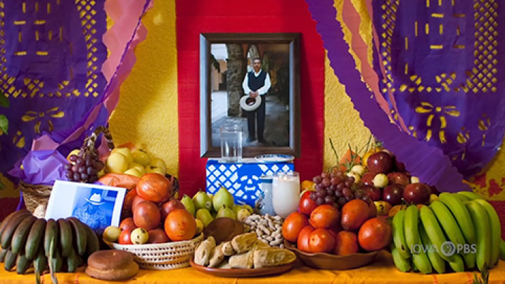 Fruits and bread sit on a table with lit candles. A photo of a man is propped on the wall behind it.