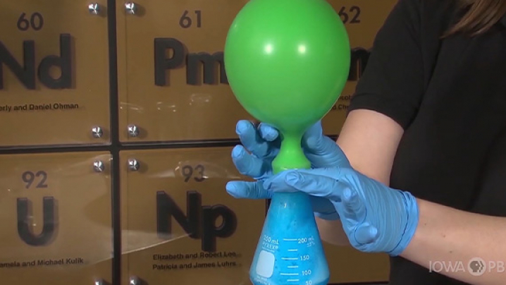 A woman holds a beaker filled with a foamy turquoise substance. A balloon is attached to the top of the beaker as it inflates.