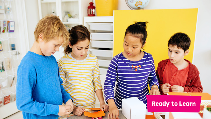Four children gathered around a table contemplating how to build a structure out of paper.