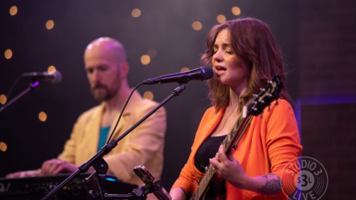 A woman in an orange top playing guitar and singing into a microphone with a man in a yellow jacket behind her on a keyboard