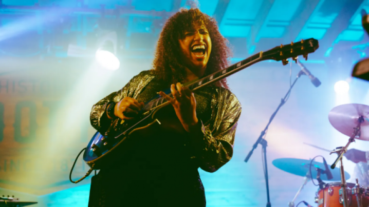 Musician energetically playing guitar on stage, lit with blue and green lights.
