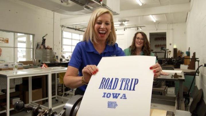 Host Brooke Kohlsdorf holding a "ROAD TRIP IOWA" poster