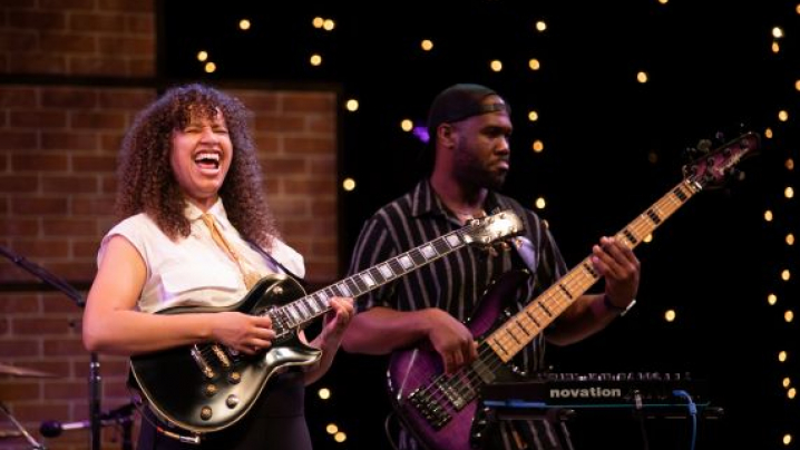 Two musicians performing on stage with decorative string lights and a brick wall background.