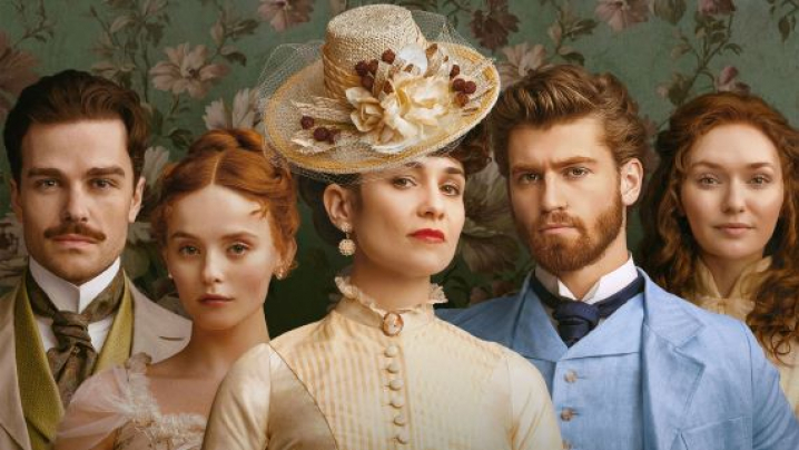 Five people in vintage clothing stand in front of floral wallpaper.
