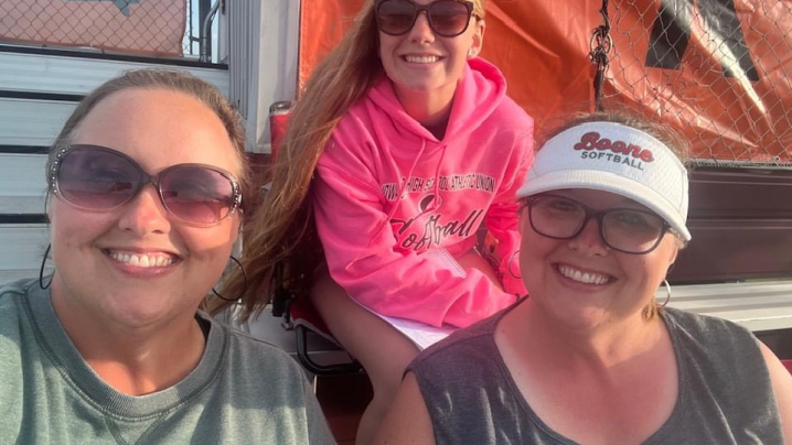 Three young women in tank tops sit in the bleachers while taking a selfie.
