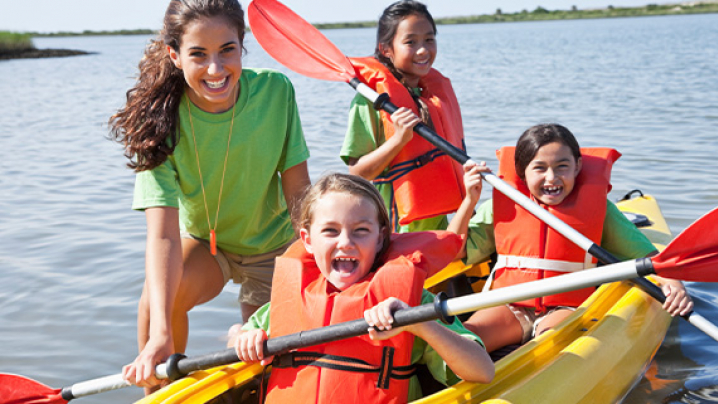 Three children in orange life jackets and an adult in a green shirt with red paddles in a yellow kayak on a lake.