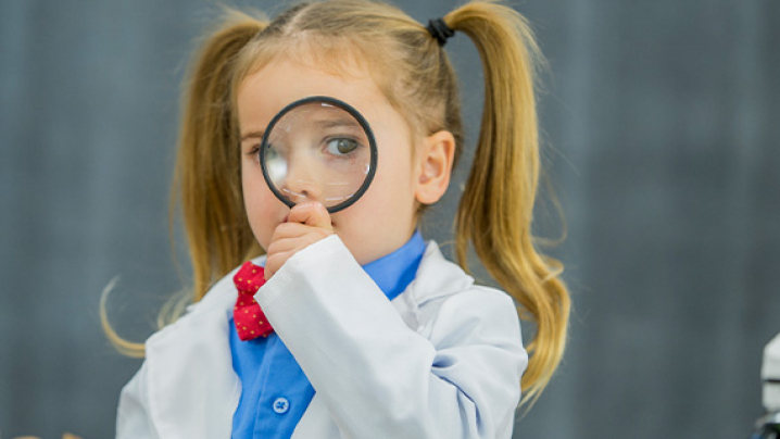 A young girl with pigtails in a white lab coat, holding a magnifying glass.