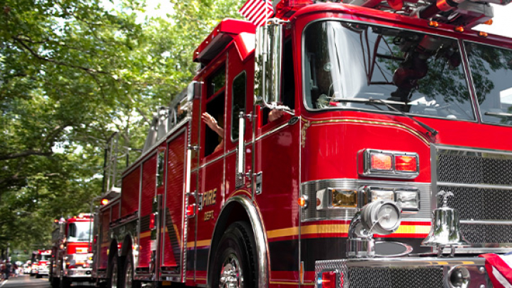 Red firetrucks driving down a tree lined street on a sunny spring day.