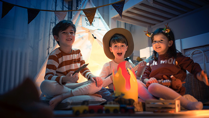 Three children sitting around a faux campfire in a cozy indoor setting, smiling and enjoying playtime together.
