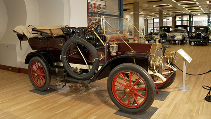 A vintage burgundy car from the early 1900s with red wooden wheels, displayed in a museum with other classic vehicles in the background.
