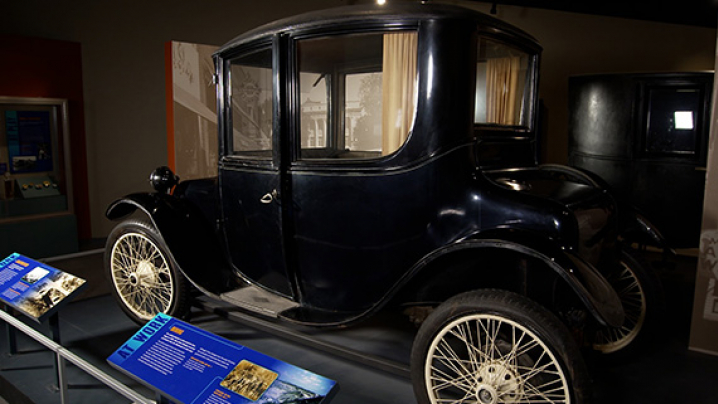 A vintage black automobile with large spoked wheels and tall windows is showcased in a museum, accompanied by informative display panels with blue backgrounds.