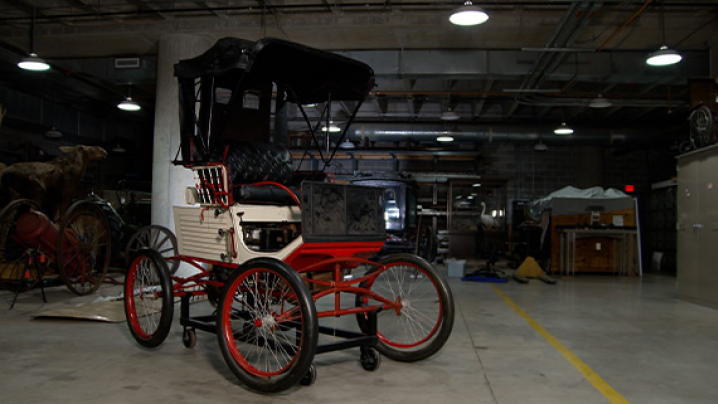 A vintage red and white carriage with a black canopy sits in a dimly lit museum storage area.