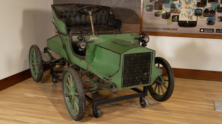A vintage green car with a tufted black leather seat and spoked wheels displayed in a museum.