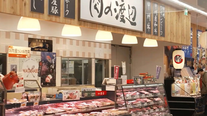 Interior of a grocery market displaying a variety of meats in a refrigerated case, with signage in Japanese and English, including the name "の渡辺."