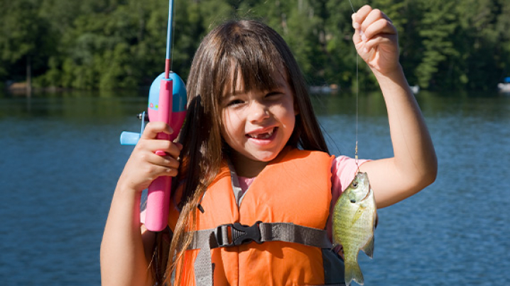 A young girl in an orange life jacket smiles while holding up a small fish caught on a fishing line near a lake.