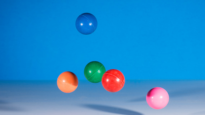 Five colorful plastic balls (blue, orange, green, red, and pink) are suspended in mid-air against a blue background.