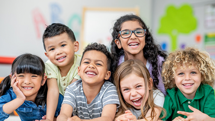 A diverse group of six smiling children sits closely together in a bright classroom, showcasing their joyful expressions.