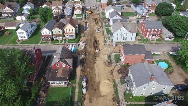 Aerial view of a construction site on a residential street, with disturbed ground and construction equipment surrounded by various houses