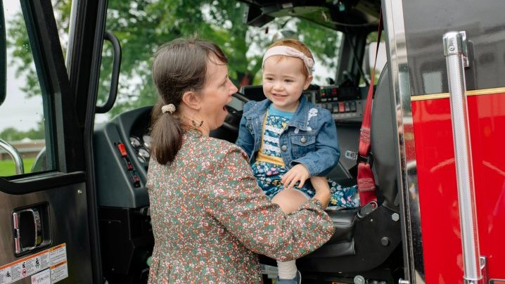 A mom holding her child as the child smiles sitting in the front seat of a fire truck.