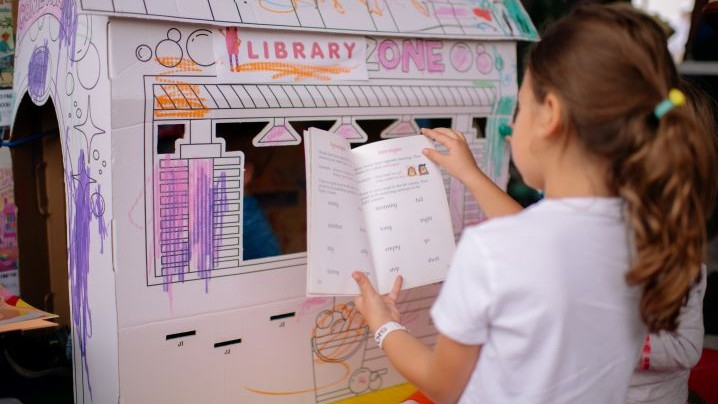 A girl reading a book in front of a cardboard house.