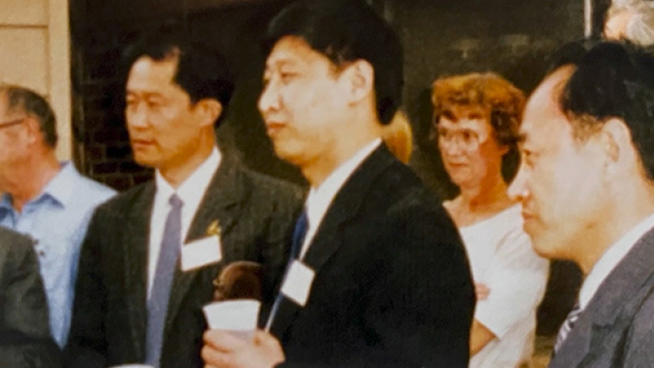 Adults in formal attire gather indoors; two central men wear name badges, and one holds a cup.