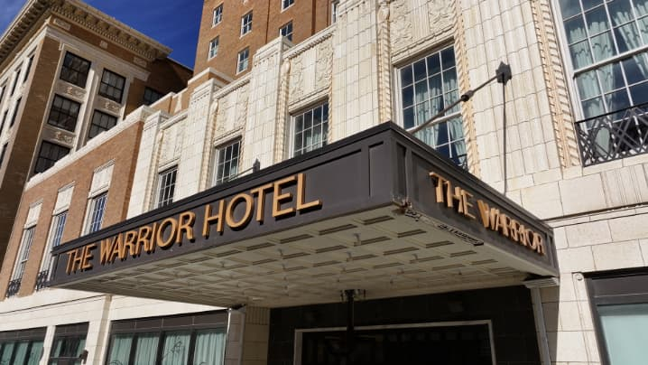 Historic hotel entrance with a black marquee reading “THE WARRIOR HOTEL” on a cream stone and brick building.