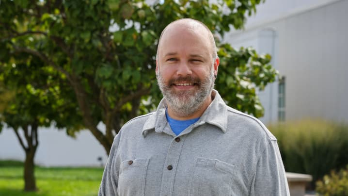 A man in a grey button up shirt standing in front of a tree. 