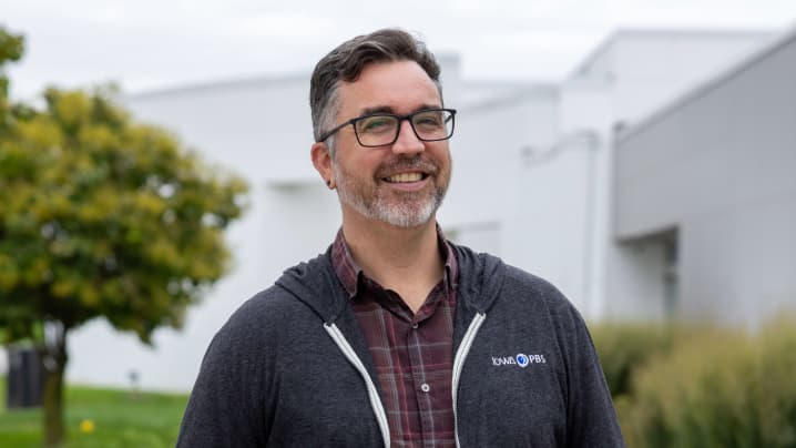 Portrait of smiling Iowa PBS producer director Patrick Boberg wearing glasses, a burgundy plaid shirt, and a dark gray Iowa PBS hoodie outdoors.