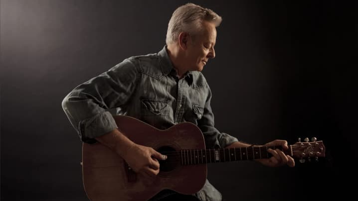 Tommy Emmanuel seated and playing a brown acoustic guitar in a dimly lit studio.