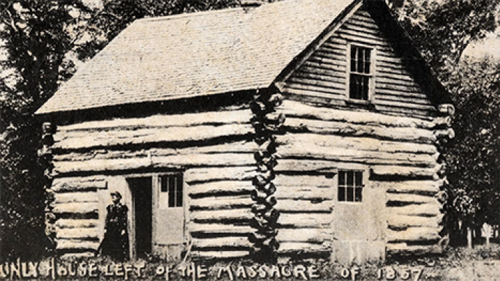 A black and white photo of a Spirit Lake Massacre log house. A woman stands outside a doorway. Text at the bottom of the photo reads, "ONLY HOUSE LEFT OF THE MASSACRE OF 1857."