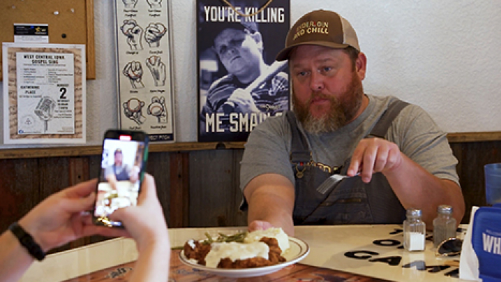 JayJay Goodvin (Iowa Gallivant) holds up a plate of food while somebody documents him and the plate with a cell phone for social media.