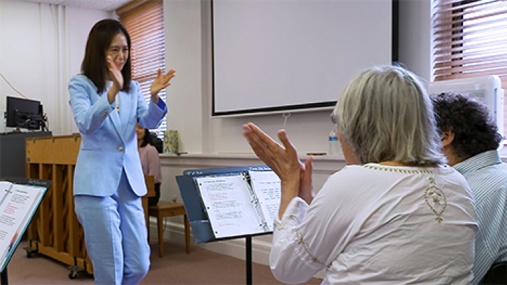 A woman musical instructor leads members of the Tremble Clefs during a song. Their hands are up and clapping.