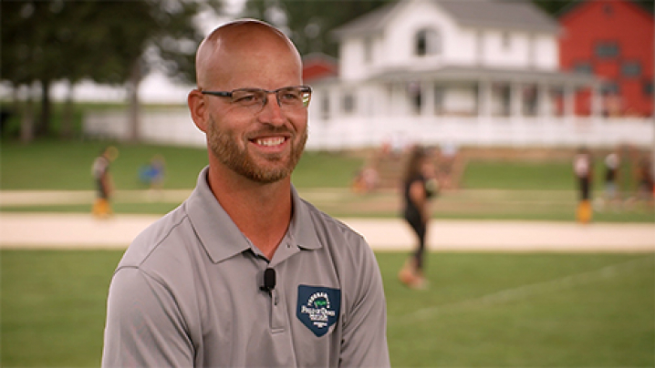 Russ Bolibaugh smiles on the Field of Dreams baseball field. The white farmhouse is over his left shoulder. People play catch in the background.