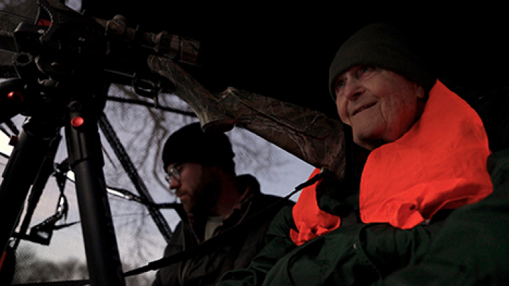 Ron Howing sits behind the scope of a shotgun with an orange safety vest on. Another man is in the background.
