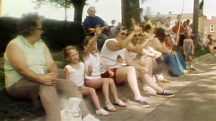 Families line a parade route - sitting on the street curb and waving to those in the parade.
