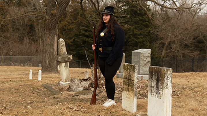 A high school student wears a civil war uniform while holding a gun next to several tombstones and tells a story about who from history she is representing.
