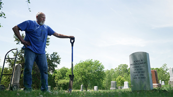 A low angle view of an older man in a blue shirt and jeans on the left standing in a graveyard with one hand on his hip and an one hand on a shovel which stands vertical. He is facing the right side of the image and focusing on a gravestone that reads, "JOHN B MONTGOMERY, GOODE'S CO, 17 US INF, WAR OF 1812, APR 1786, MAY 29 1880."