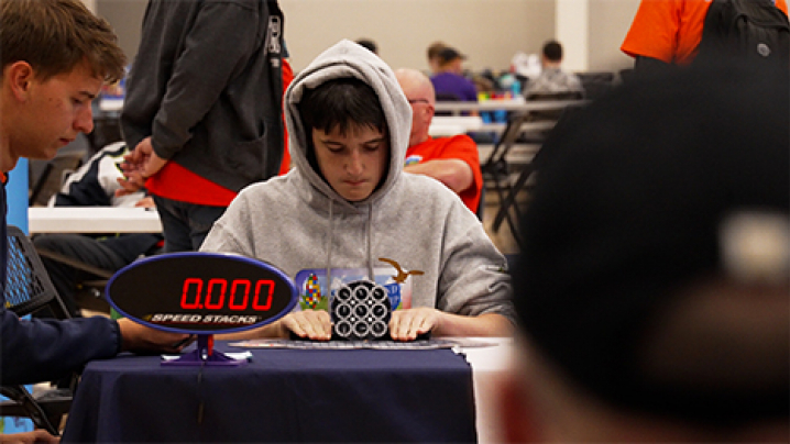 Two boys at a speedcubing competition table. An oval timer on the table reads 0.000. Other participants and officials standing and sitting at tables behind them. View is partially obstructed by the blurred view of the back of a man's head wearing a black hat.