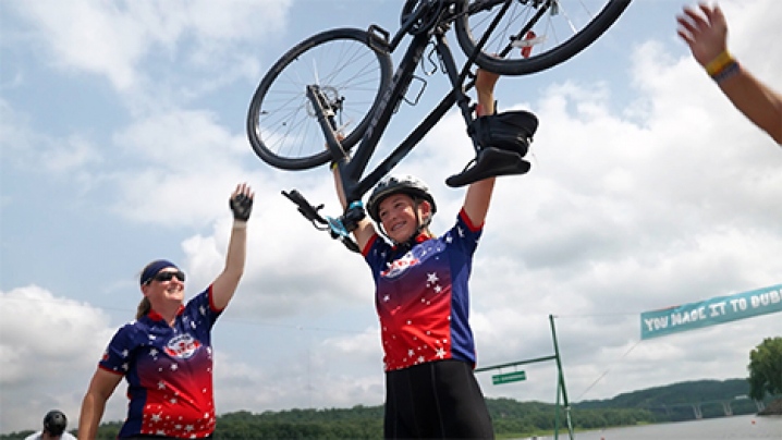 A member of the Dream Team cycling team raises their bike above their head in celebration at the Mississippi River. Teammates flank their side.