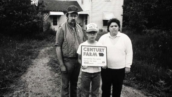 Family stands in front of farm home holding a century farm sign during the farm crisis