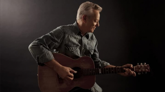 Tommy Emmanuel seated and playing a brown acoustic guitar in a dimly lit studio.