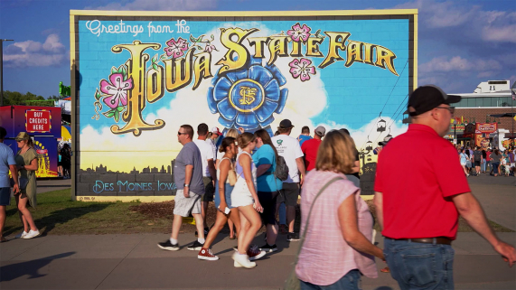 Fairgoers walk past a large mural that says, "Iowa State Fair" and has a blue ribbon on it.