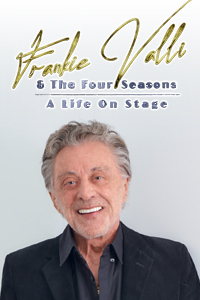 Frankie Valli smiling at the camera in a gray unbuttoned at the neck shirt and jacket.