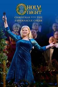 Star and performer, Megan Hilty stands smiling onstage in front of The Tabernacle Choir with her arms open - she is holding a microphone up in the air with her right hand.