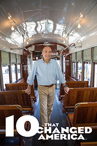 A white 10 That Changed America logo underneath host, Geoffrey Baer who stands smiling in the aisle an empty public transport street car with wooden seats.