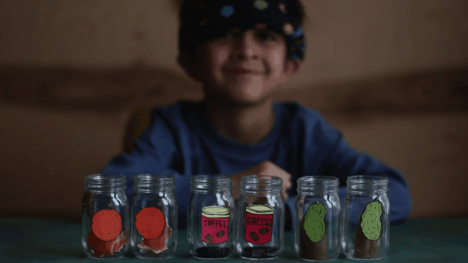 Six jars in a row in front of a young child. From left to right there are two jars with a drawing of an orange on them, two jars with text on them and two jars with a drawing of a potato on them.
