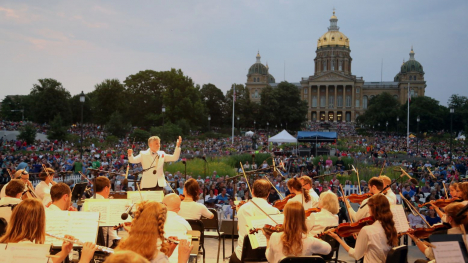 In the foreground, the Des Moines Symphony wearing white shirts plays at Yankee Doodle Pops. The Iowa Capitol and a crowd on its lawn are in the background.