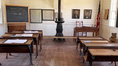 The inside of a one-room schoolhouse, which contains a wood burning stove, chalkboard, and desks. 